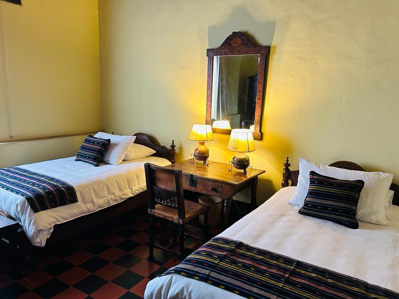 Une chambre avec deux lits, un bureau en bois, deux lampes et un miroir au mur.