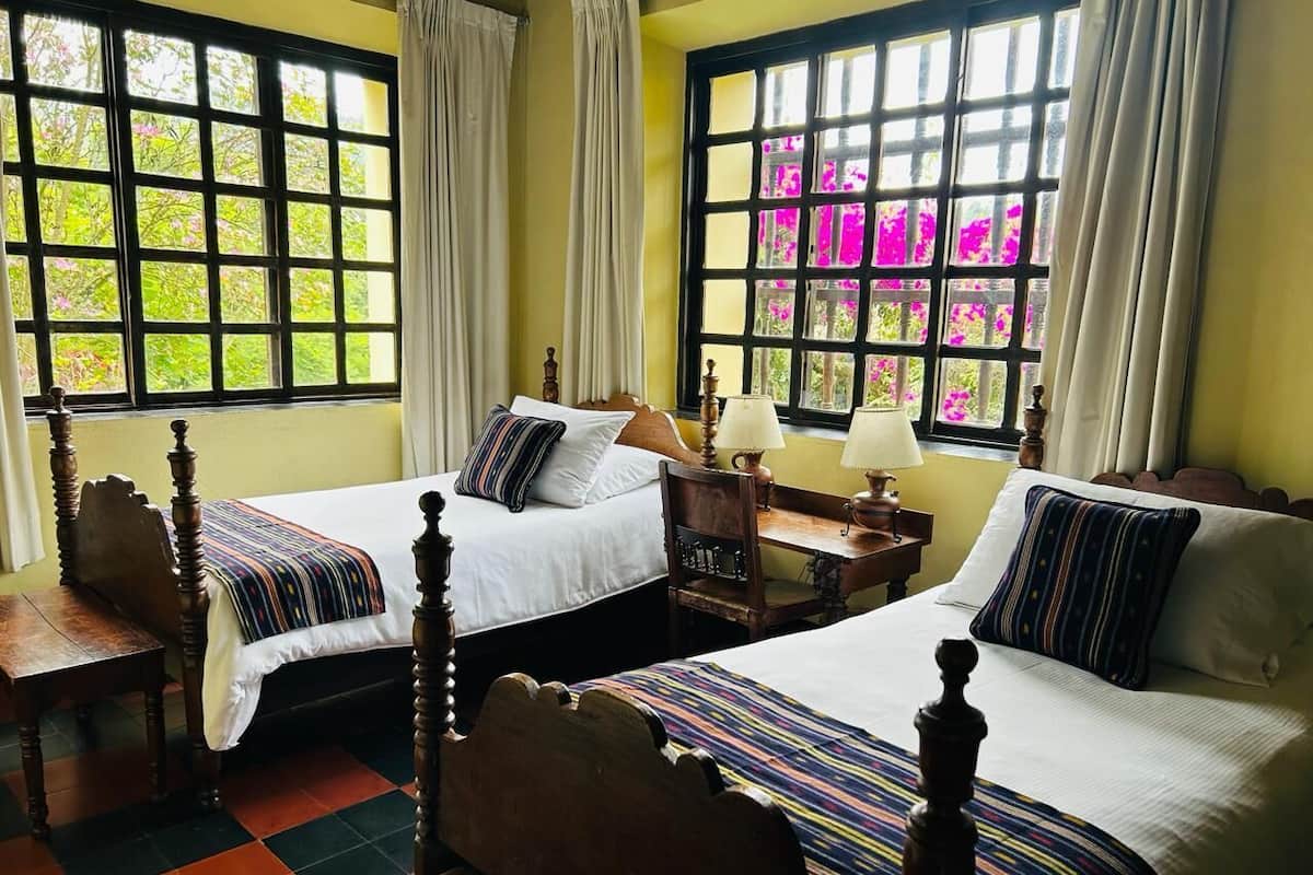 A room with two beds, a wooden chair, a nightstand, and a window with curtains.