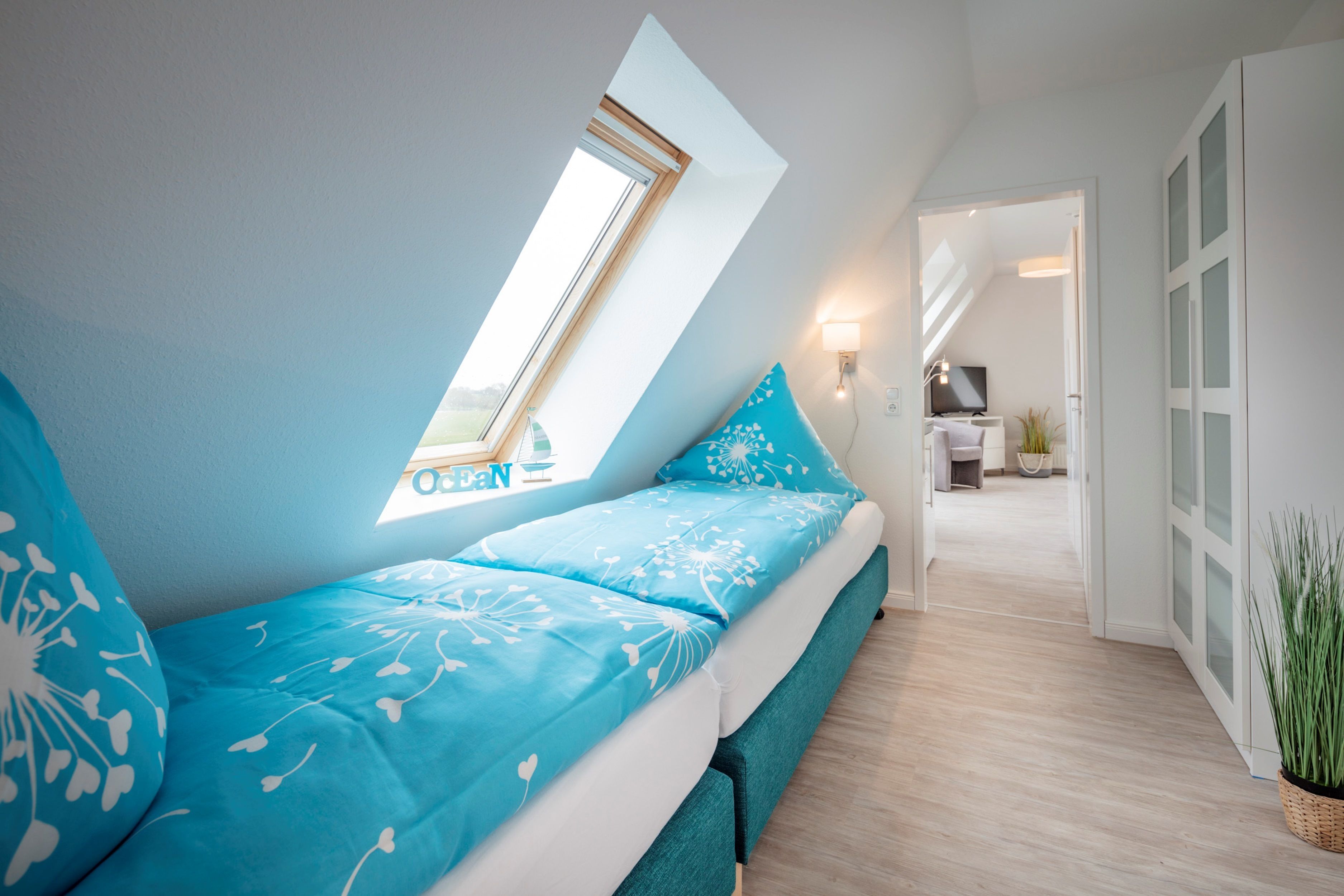 A bedroom with a skylight, a bed with blue bedding, a bedside table, and a view into the hallway.