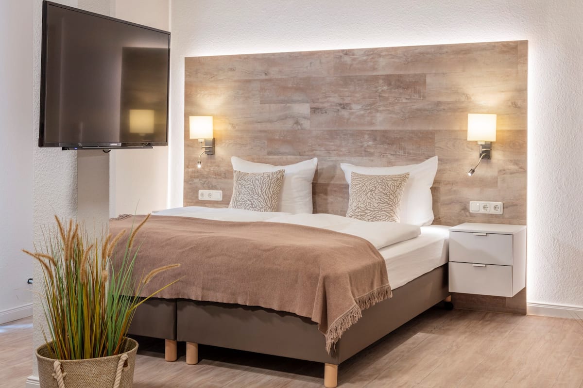 A modern bedroom with a wooden headboard, a flat-screen TV mounted on the wall, bedside lamps, and a bedside table with a lamp.