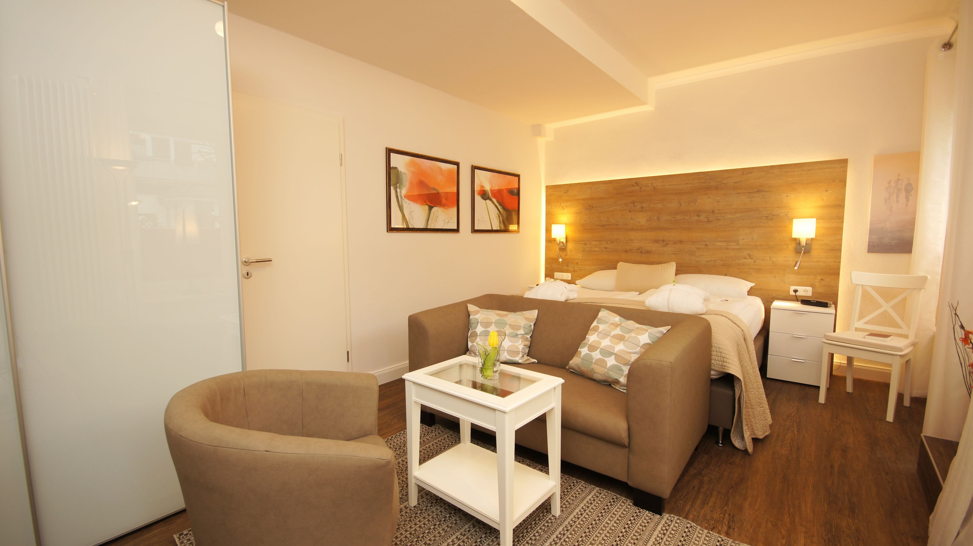 Ein modernes Hotelzimmer mit einem braunen Sofa, einem kleinen weißen Tisch mit einer Vase, einem hölzernen Kopfteil, einem Nachttisch mit einer Lampe und einem weißen Sessel.