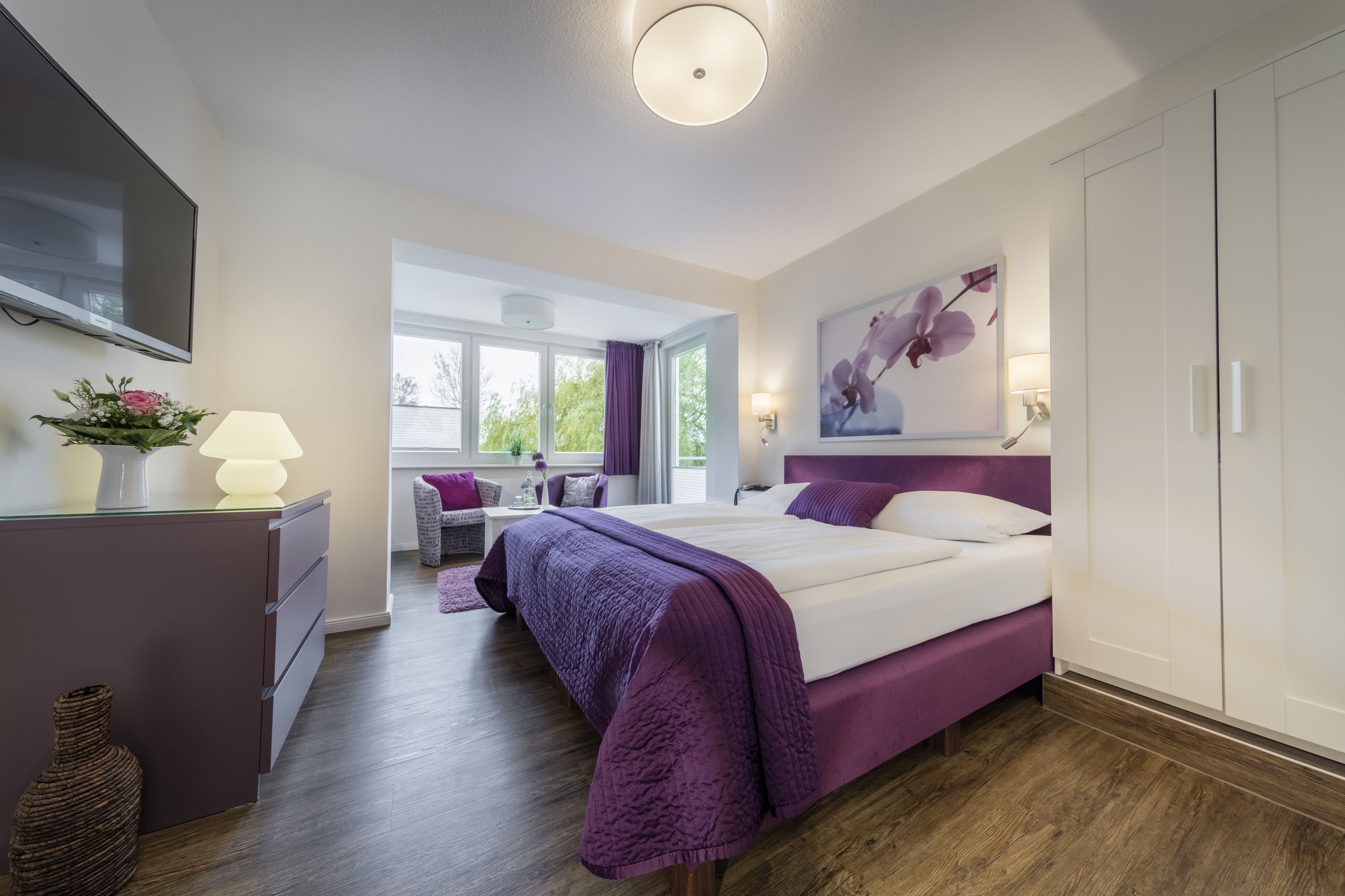 Un dormitorio moderno con una cama grande, un escritorio y un televisor. Hay una ventana con cortinas y un cuadro en la pared.