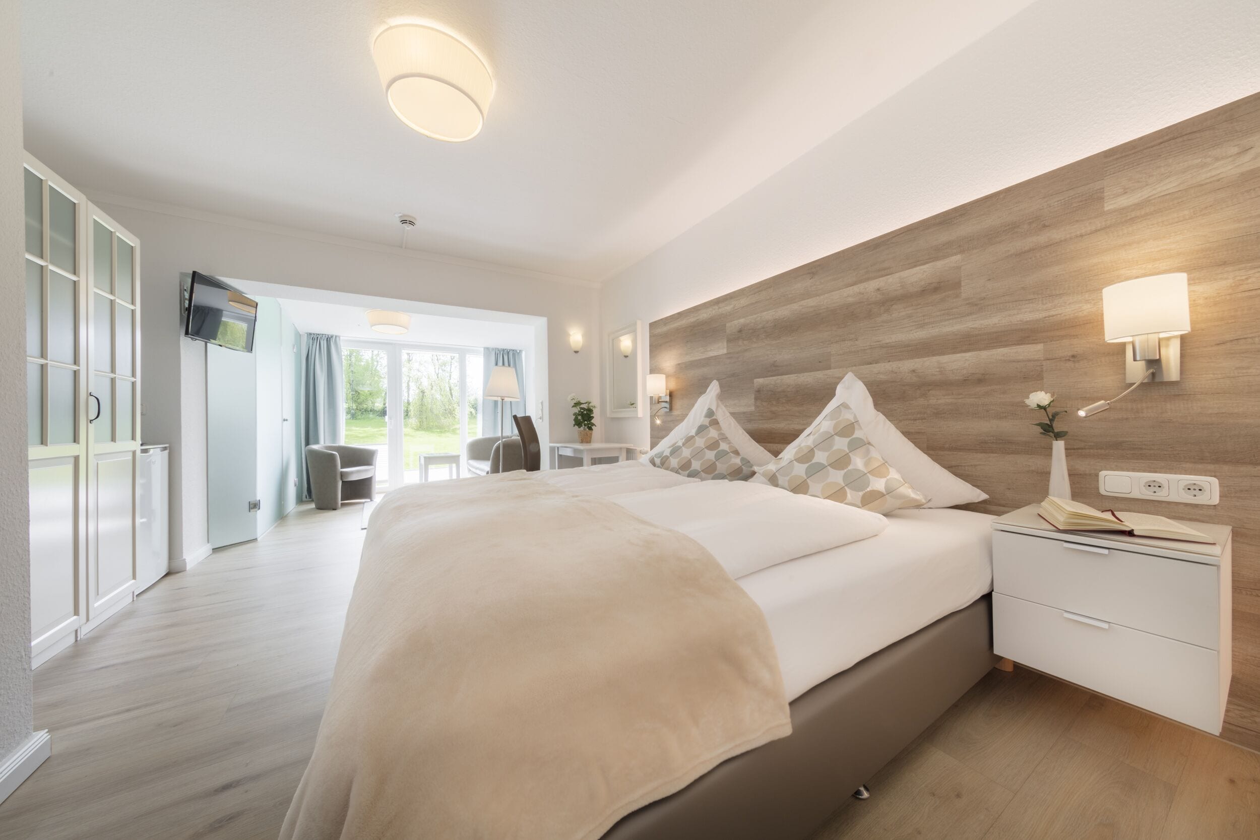 A modern hotel room with a large bed, a wooden accent wall, a bedside table with a lamp, and a view of the outdoor area.