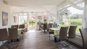 A dining area with tables and chairs, large windows with curtains, and a view of greenery outside.