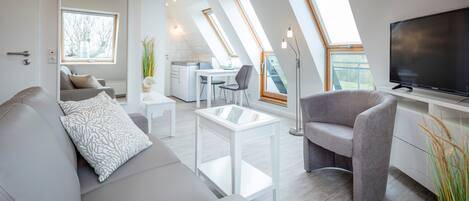 A modern living room with a sofa, a coffee table, and a TV. There are skylights and a kitchen in the background.