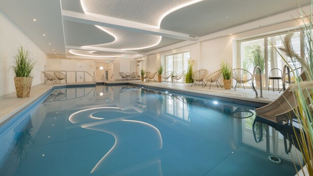 Una piscina interior con diseño moderno, que incluye un techo curvo y zonas de descanso con sillas.