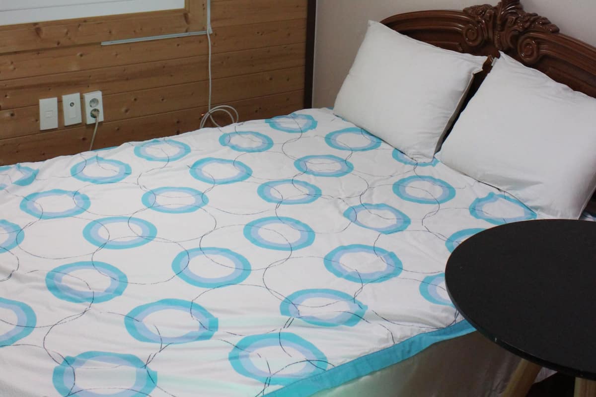 A bedroom with a wooden headboard, a window with blinds, a bed with a patterned bedspread, and a small round table.