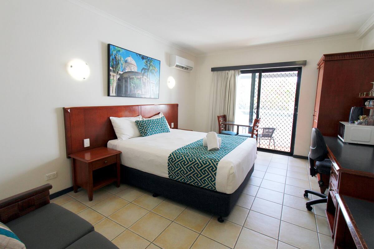 A hotel room with a bed, a desk, a chair, a small table, and a view of the outside through a sliding door.