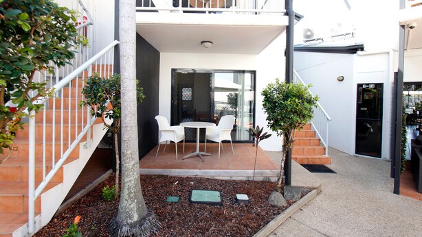 A courtyard with a patio, white chairs, a small table, and a staircase leading to an upper level. - Coffee House Apartment Motel (Rockhampton)
