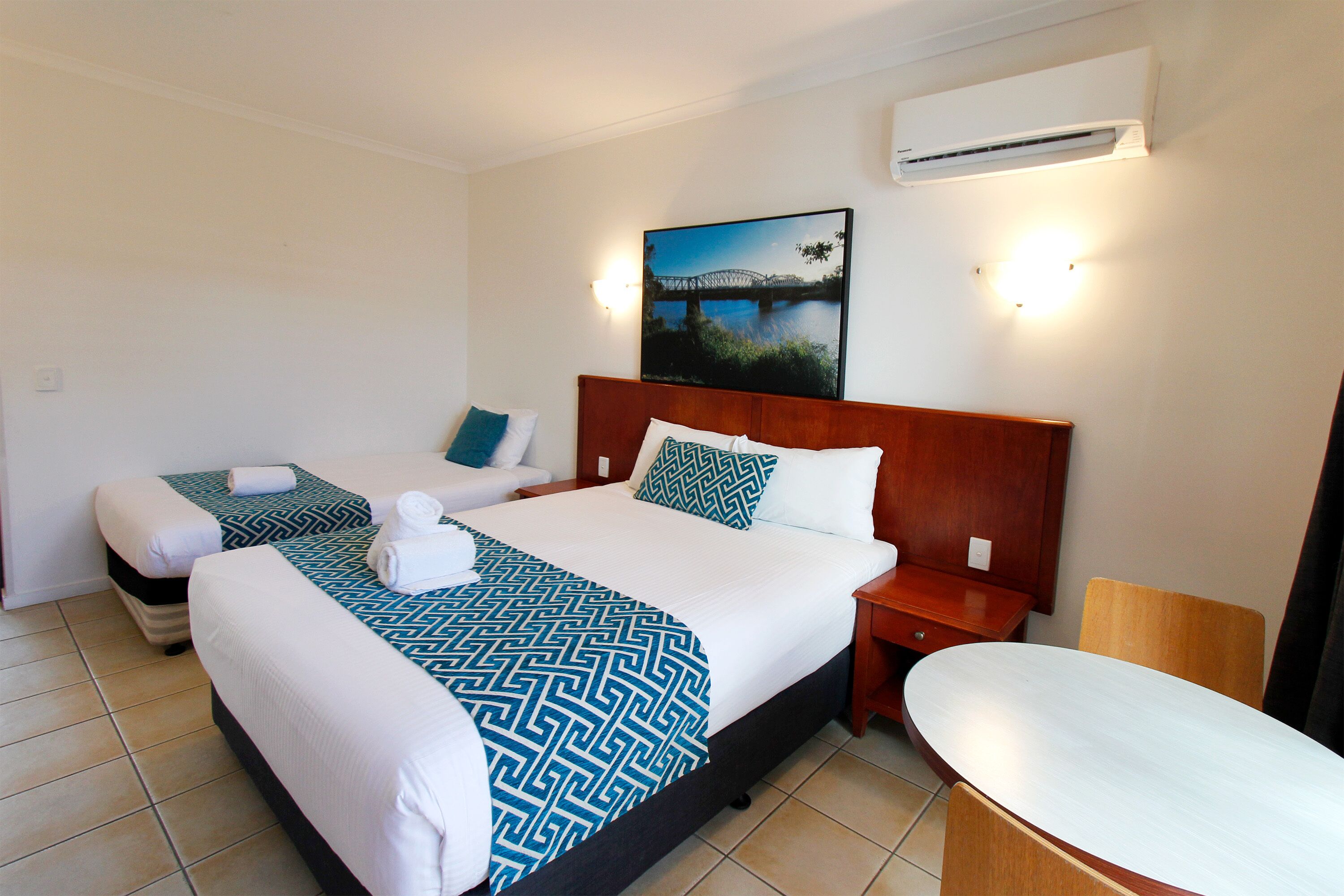 Habitación de hotel con dos camas, un cuadro grande de un puente, aire acondicionado y una mesita.