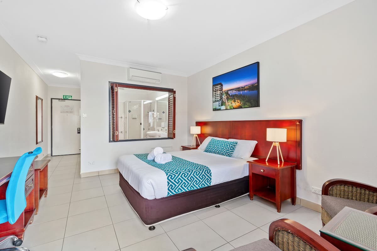 A hotel room with a bed, bedside table, lamp, chair, desk, and a large window.