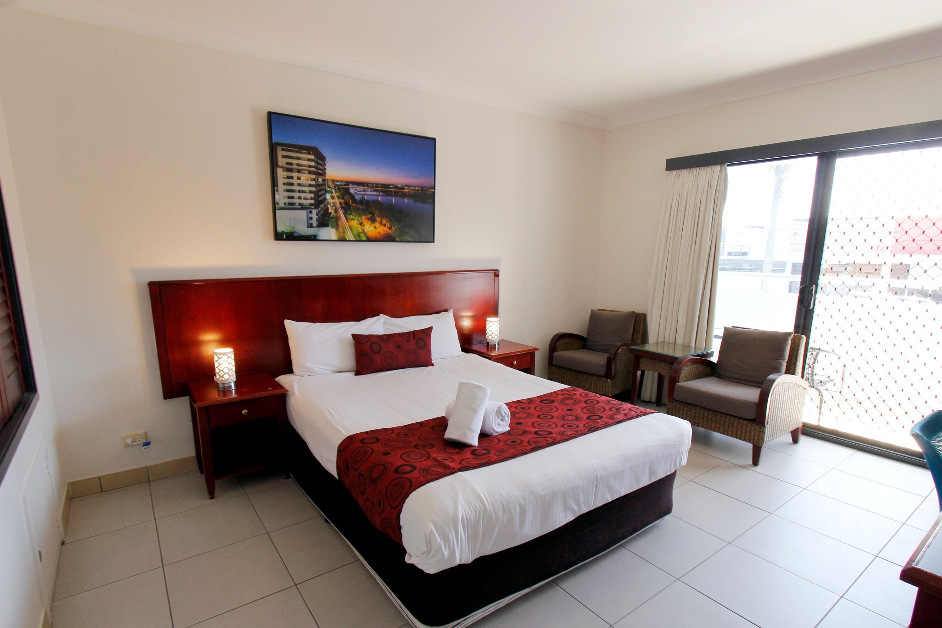 Habitación de hotel con una cama grande, una mesita de noche con lámpara, una silla y vistas a la ciudad a través de un ventanal amplio.