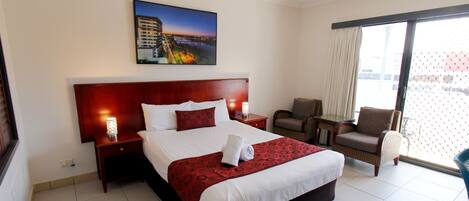 Habitación de hotel con una cama grande, una mesita de noche con lámpara, una silla y vistas a la ciudad a través de un ventanal amplio.