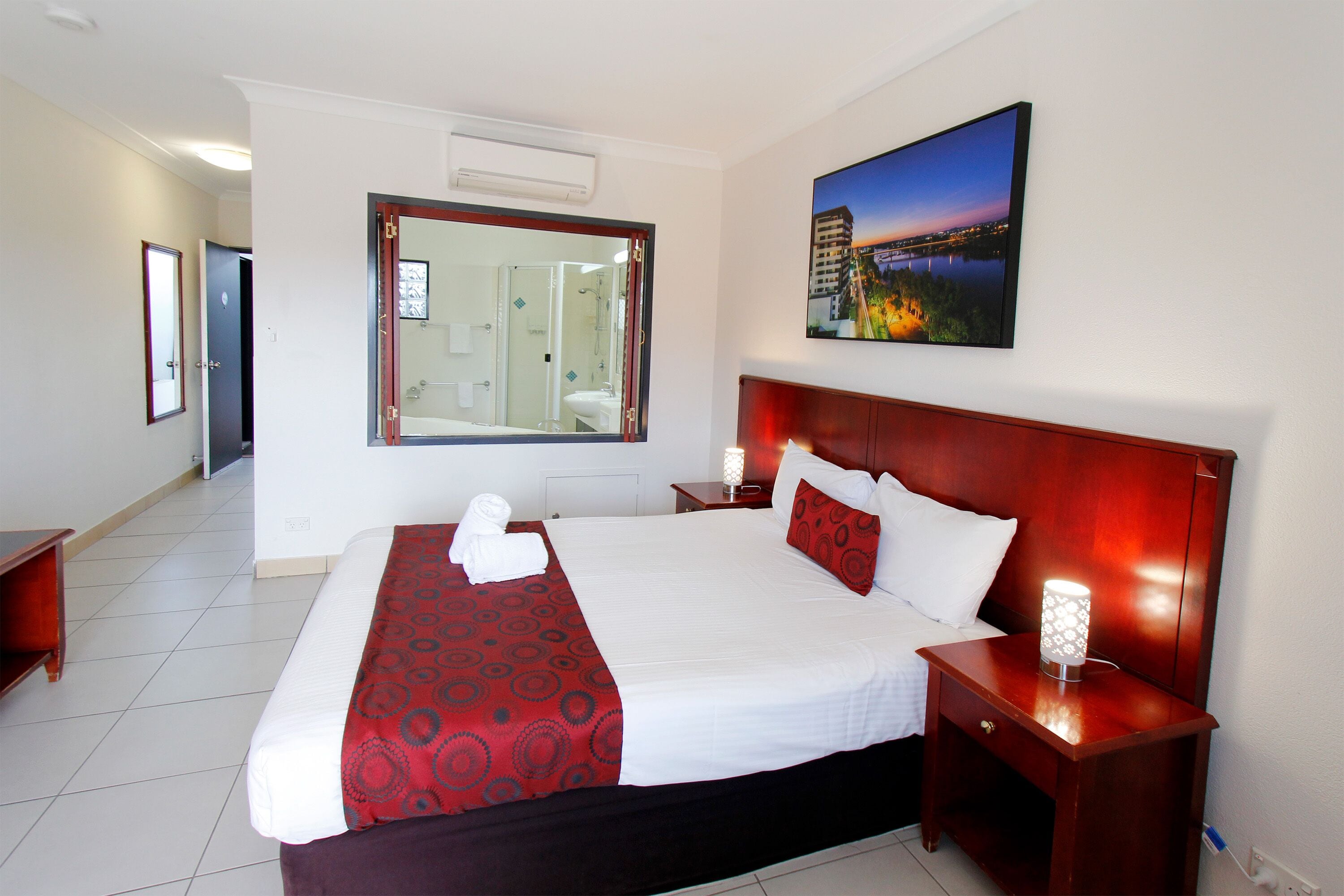 Habitación de hotel con una cama grande, una mesita de noche con lámpara, un espejo y un cuadro en la pared.