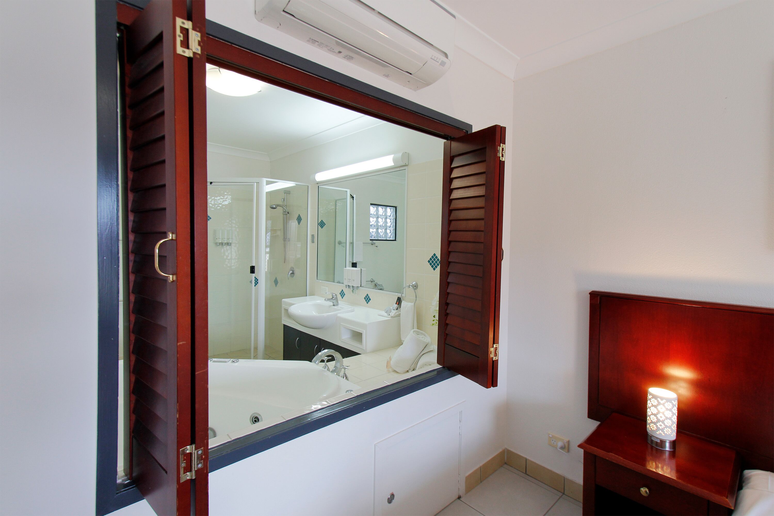 Un baño con un espejo grande, una bañera, dos lavamanos y una mesita de noche con una lámpara.