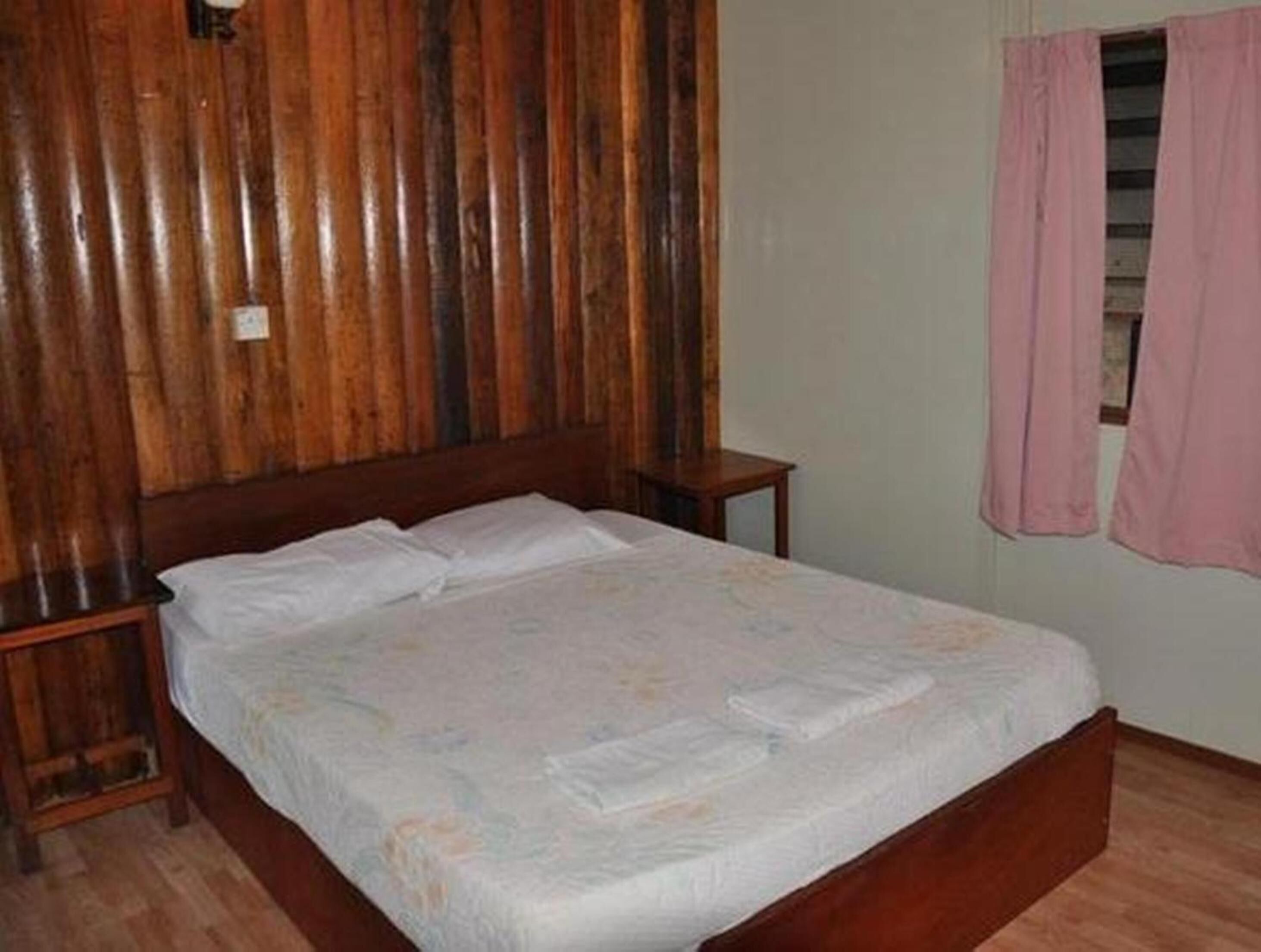 Une chambre à coucher avec une tête de lit en bois, un lit simple recouvert d’une courtepointe blanche, une table de chevet et un rideau rose.