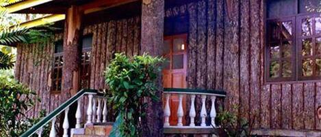 Un edificio de madera con base de piedra, una escalera con balaustres blancos y una ventana con cortinas.