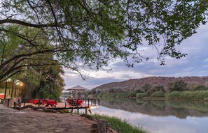 An outdoor dining area with tables and chairs, a covered seating area, and a view of a calm river with hills in the background. - Kunene River Lodge (Ruacana)