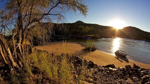 A riverbank with a boat, trees, and a clear sky. - Kunene River Lodge (Ruacana)