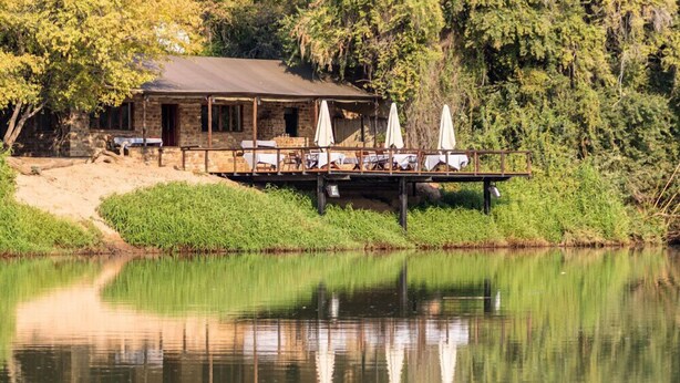A rustic stone building with outdoor seating and umbrellas by a calm lake, surrounded by greenery. - Kunene River Lodge (Ruacana)