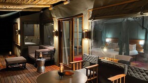 A cozy room with a wooden floor, a round table with a candle, a cushioned bench, and a bed with a canopy. - Ndhovu Safari Lodge - Campground (Mahango)