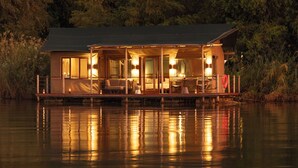 A waterfront cabin with a covered deck, large windows, and outdoor seating. - Ndhovu Safari Lodge - Campground (Mahango)