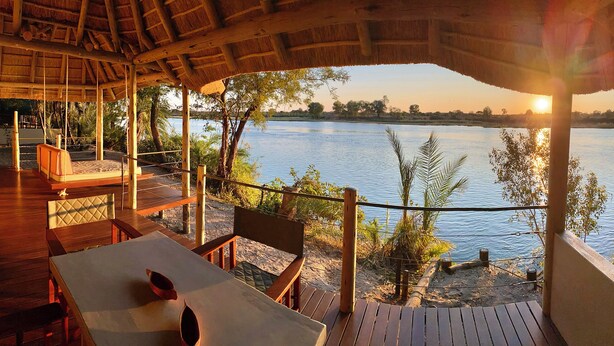 A wooden deck with a thatched roof overlooking a calm lake, featuring seating and tables. - Ndhovu Safari Lodge - Campground (Mahango)