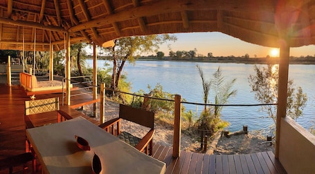 A wooden deck with a thatched roof overlooking a calm lake, featuring seating and tables.