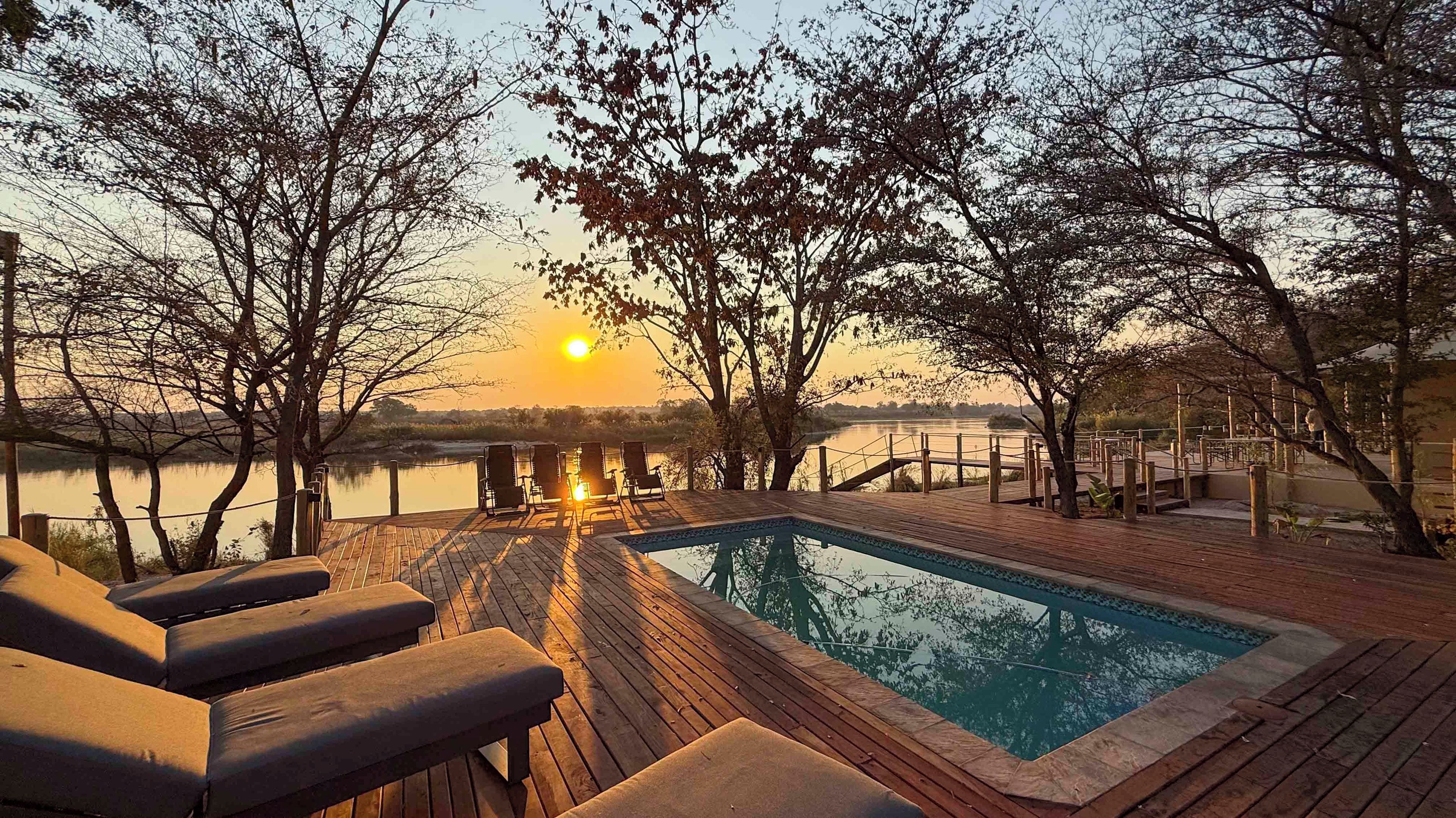 A wooden deck with a pool, lounge chairs, and a view of a sunset over a body of water.