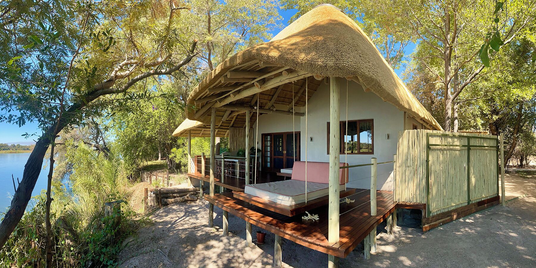 A thatched-roof bungalow with a wooden deck, a single bed, and a view of a lake.