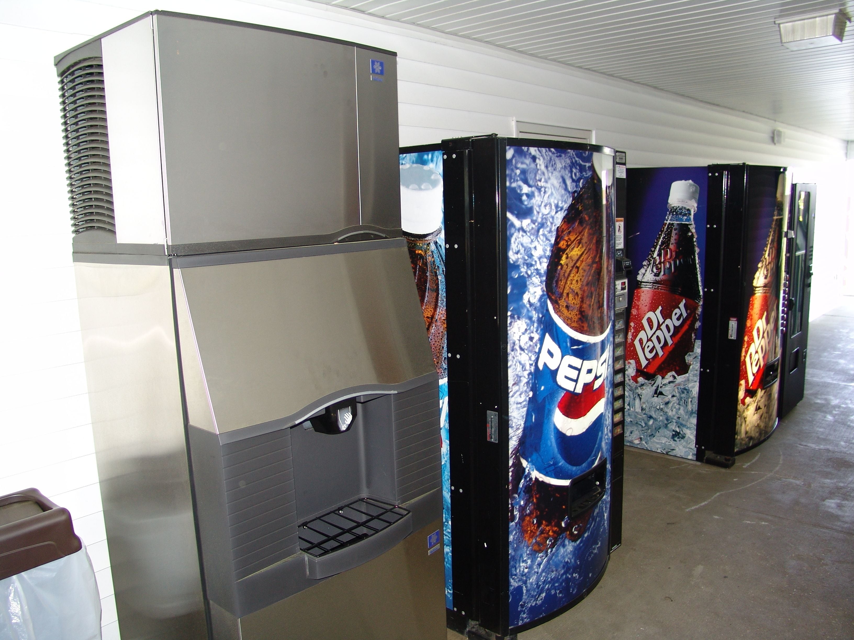 Una fila de máquinas expendedoras con logotipos de Pepsi y Dr Pepper, y un refrigerador industrial grande.