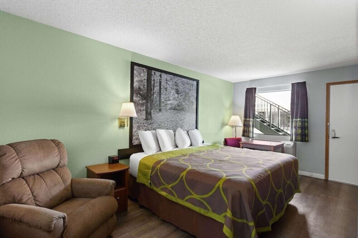 A hotel room with a bed, a chair, a nightstand, a window with curtains, and a staircase.