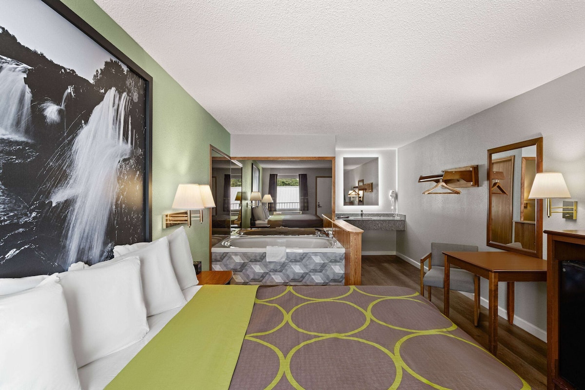 A hotel room with a large bed, a sitting area, a bathroom with a bathtub, and a large wall-mounted mirror.