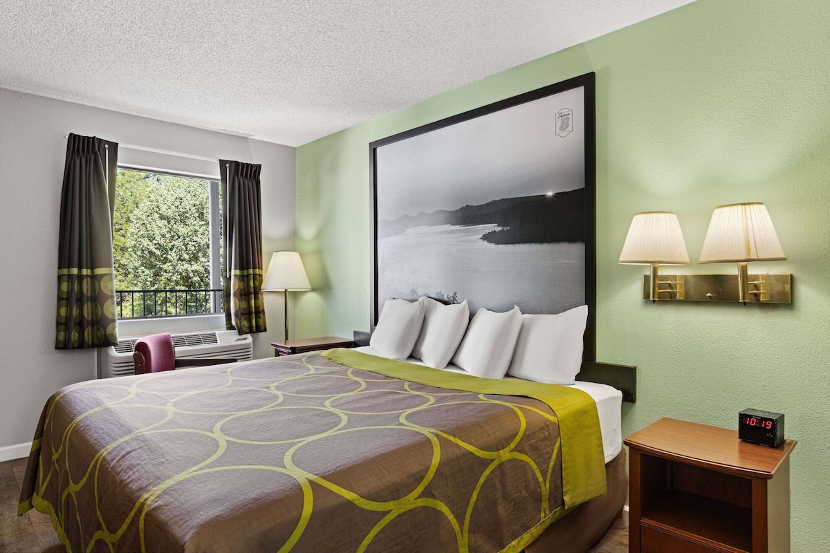 A hotel room with a large bed, a window with curtains, a nightstand with a lamp, and a wall-mounted clock.