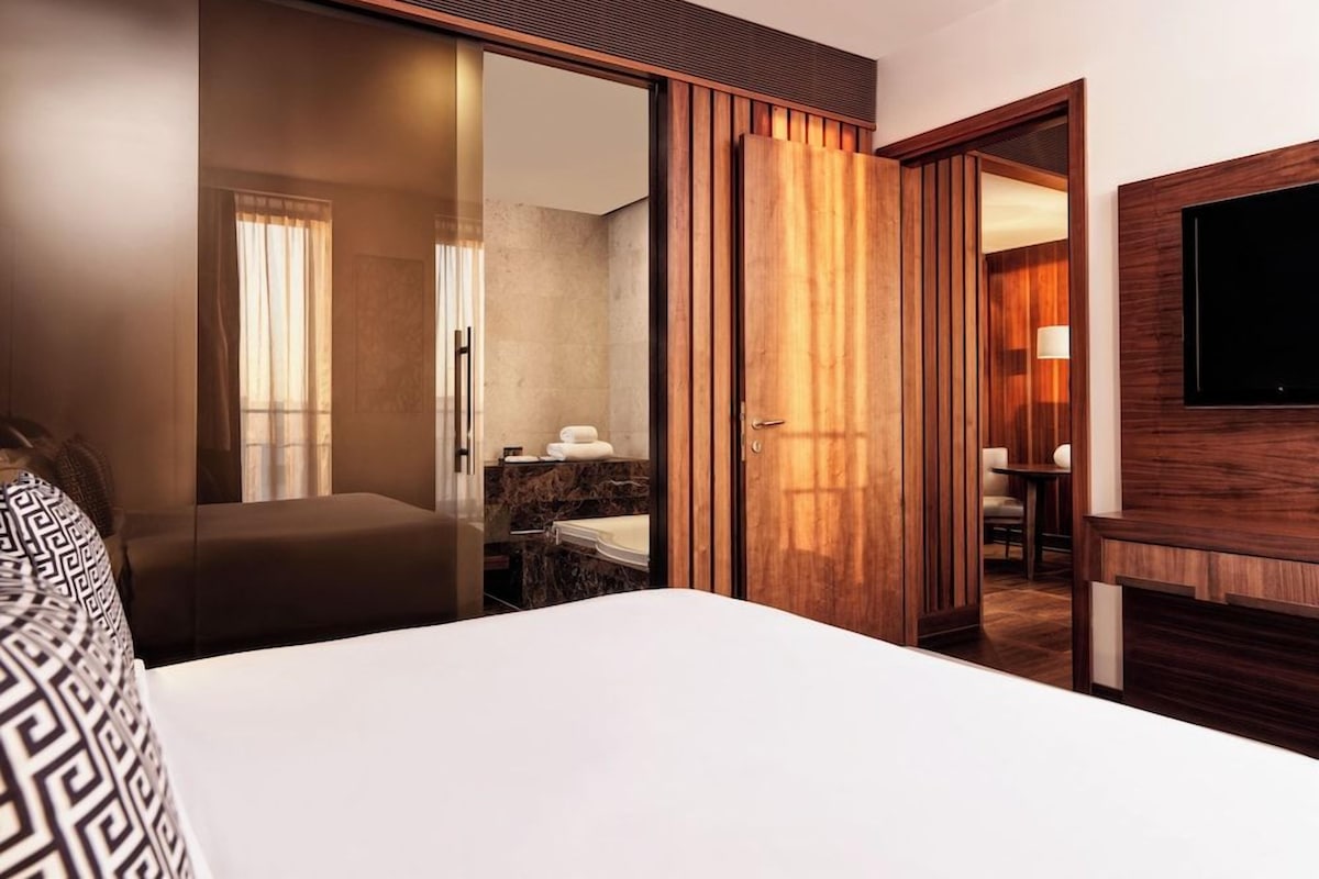 A hotel room with a bed, a desk, a TV, a bathroom with a mirror, and a sliding door.