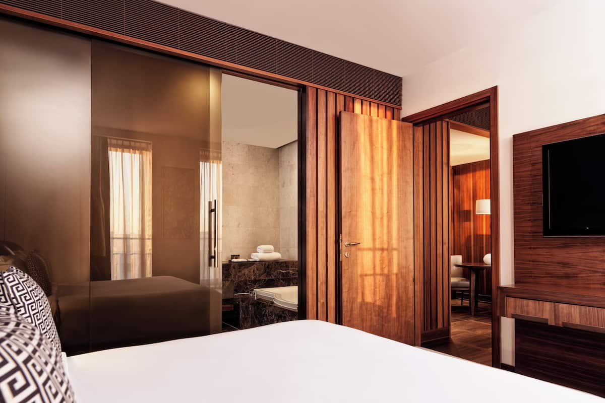 A hotel room with a bed, a bathroom with a mirror, a wooden wardrobe, and a TV mounted on the wall.