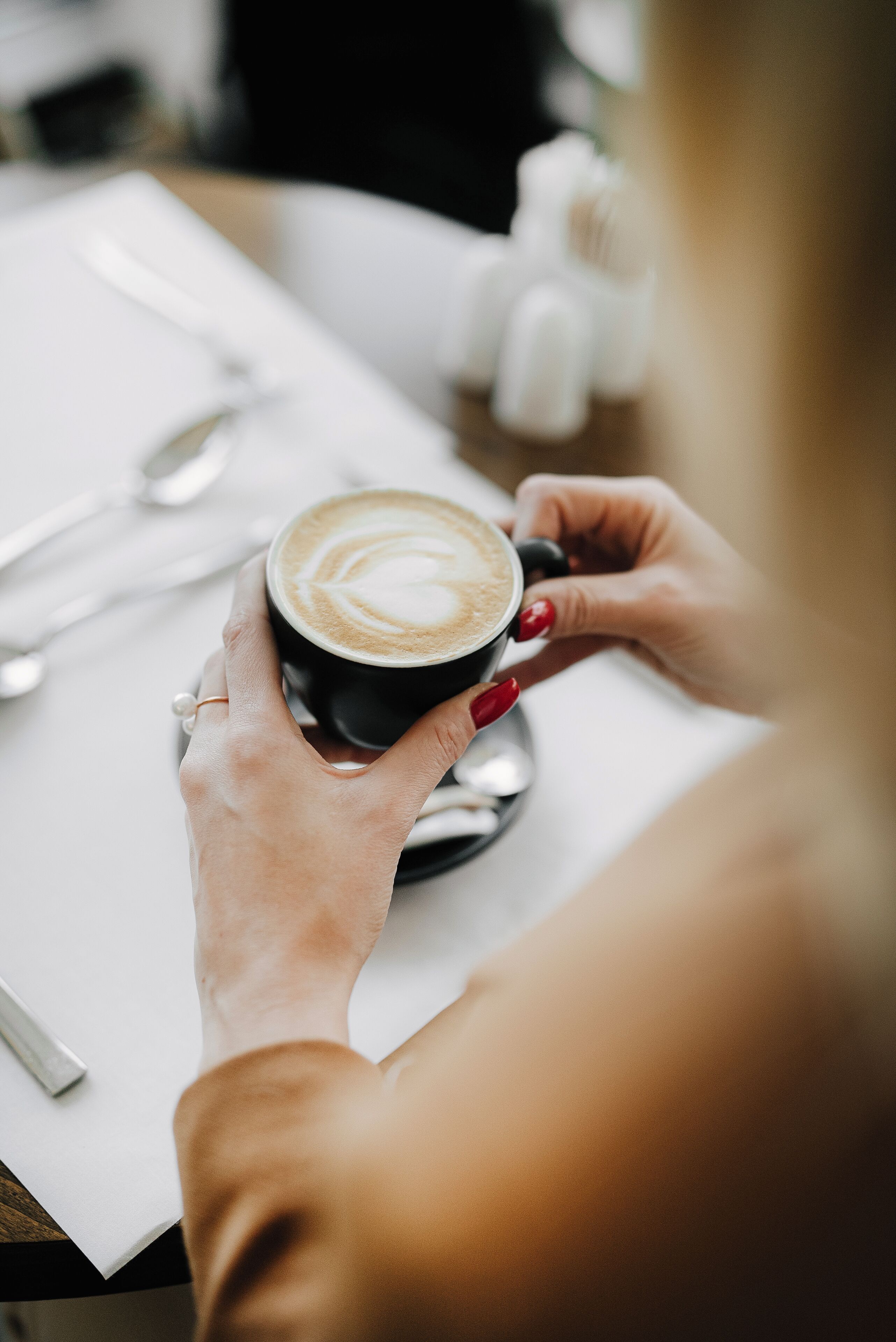 A person holding a cup of coffee with a latte art design, a spoon, and a small bottle on a white table.