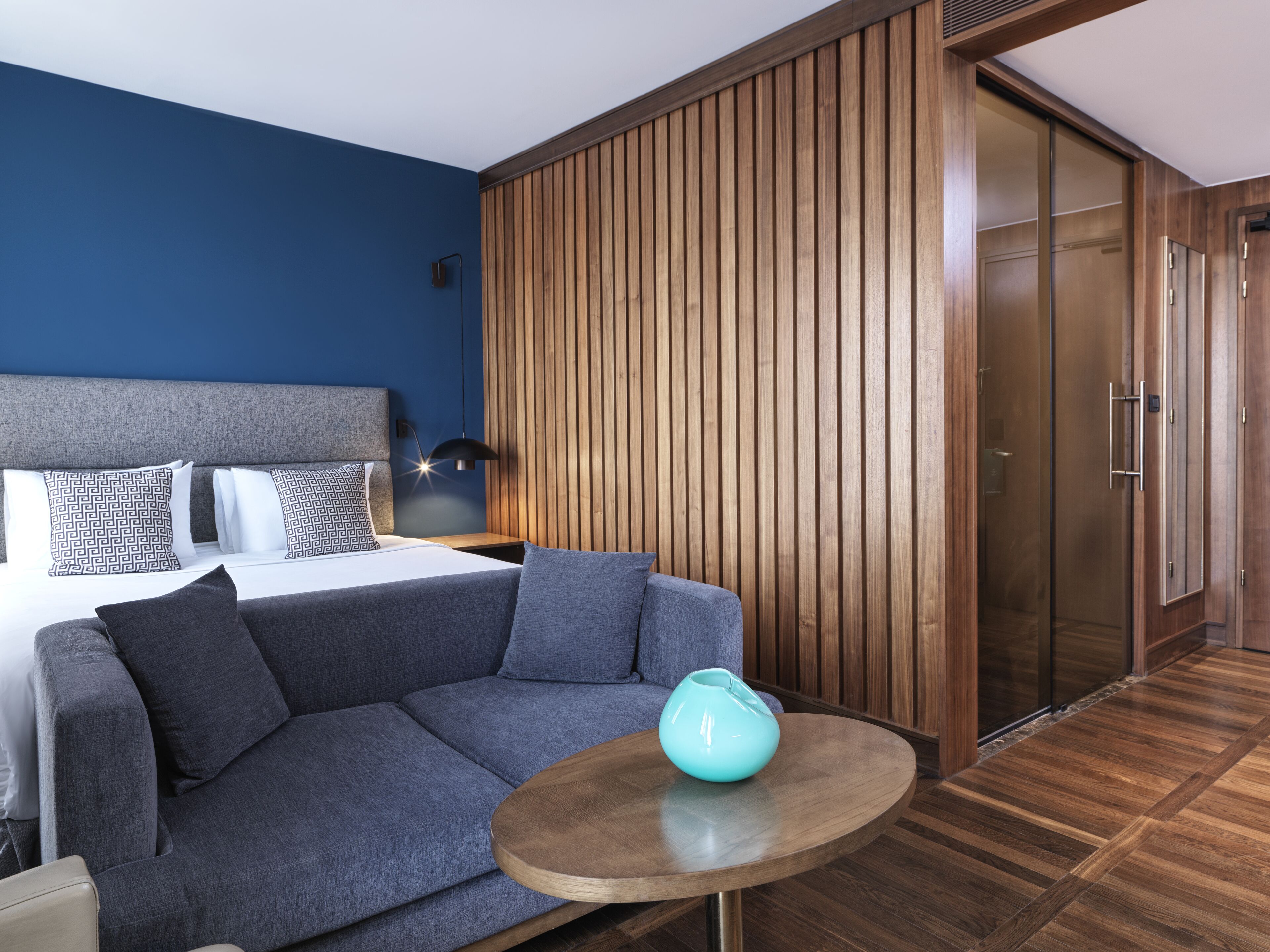 A modern hotel room with a bed, a gray sofa, a small wooden table, and a wooden panel wall.