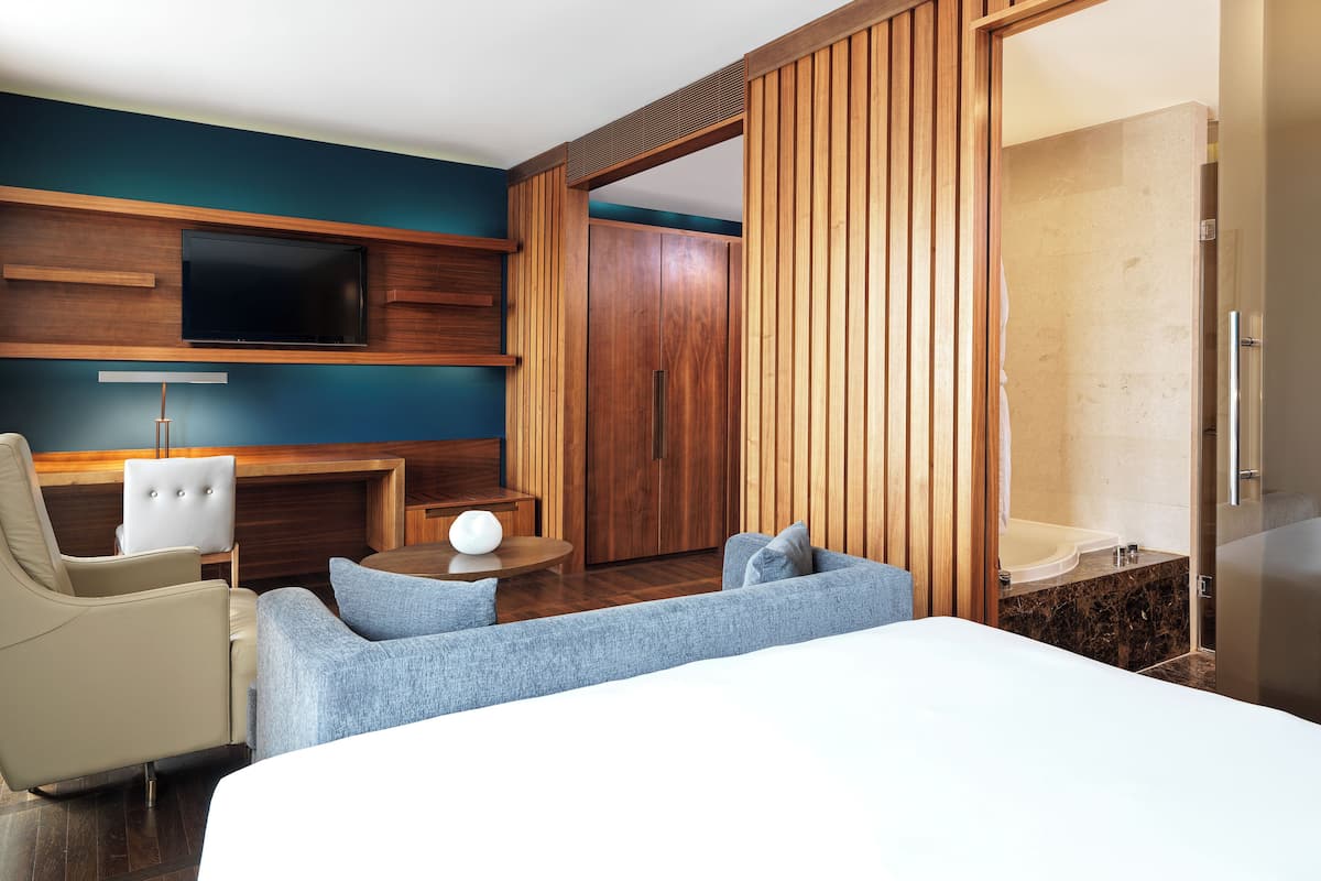 A modern hotel room with a bed, a seating area with a chair and a small table, a television mounted on the wall, and a sliding door leading to a bathroom.