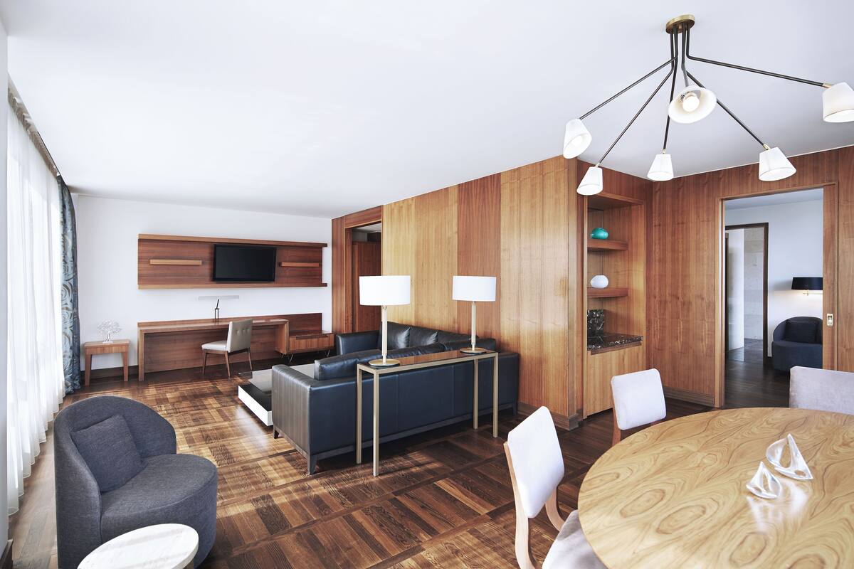A modern hotel room with a wooden floor, a sofa, a dining table, and a TV mounted on the wall.