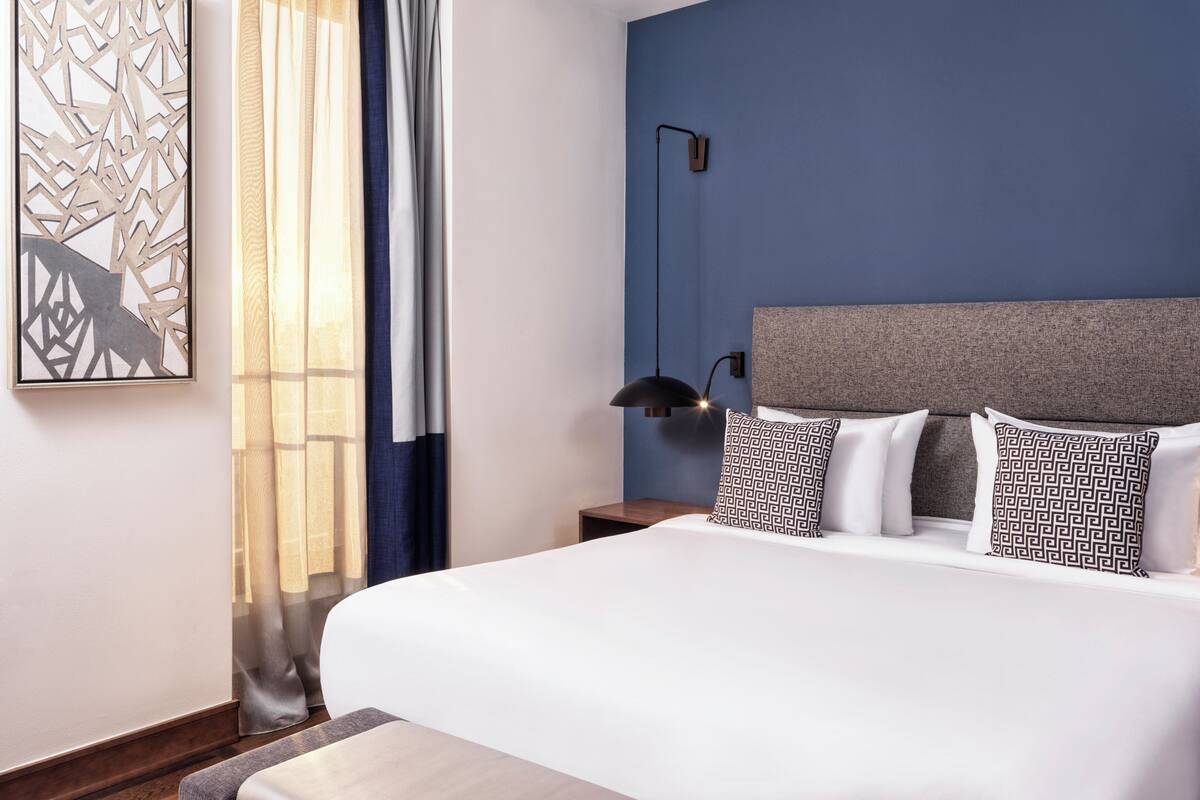 A modern hotel room with a large bed, a nightstand, a wall-mounted lamp, and a decorative piece on the wall.