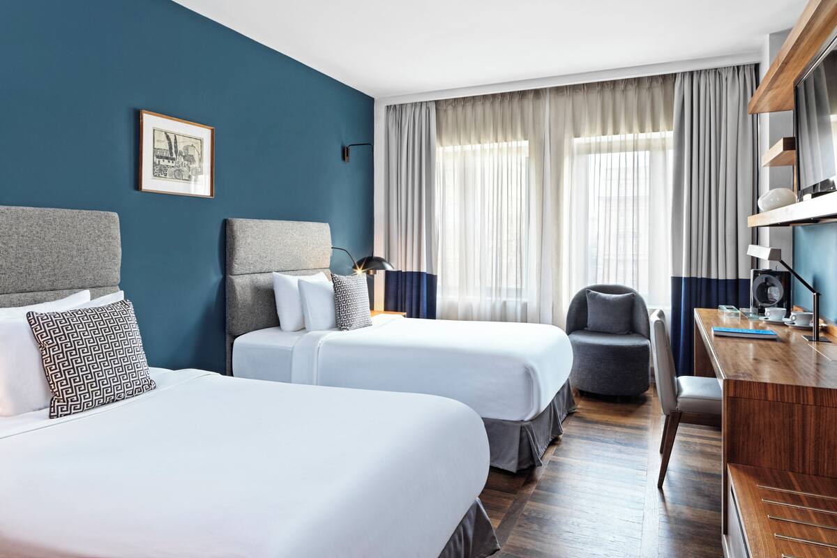 A hotel room with two beds, a desk, a chair, and a large window with curtains.