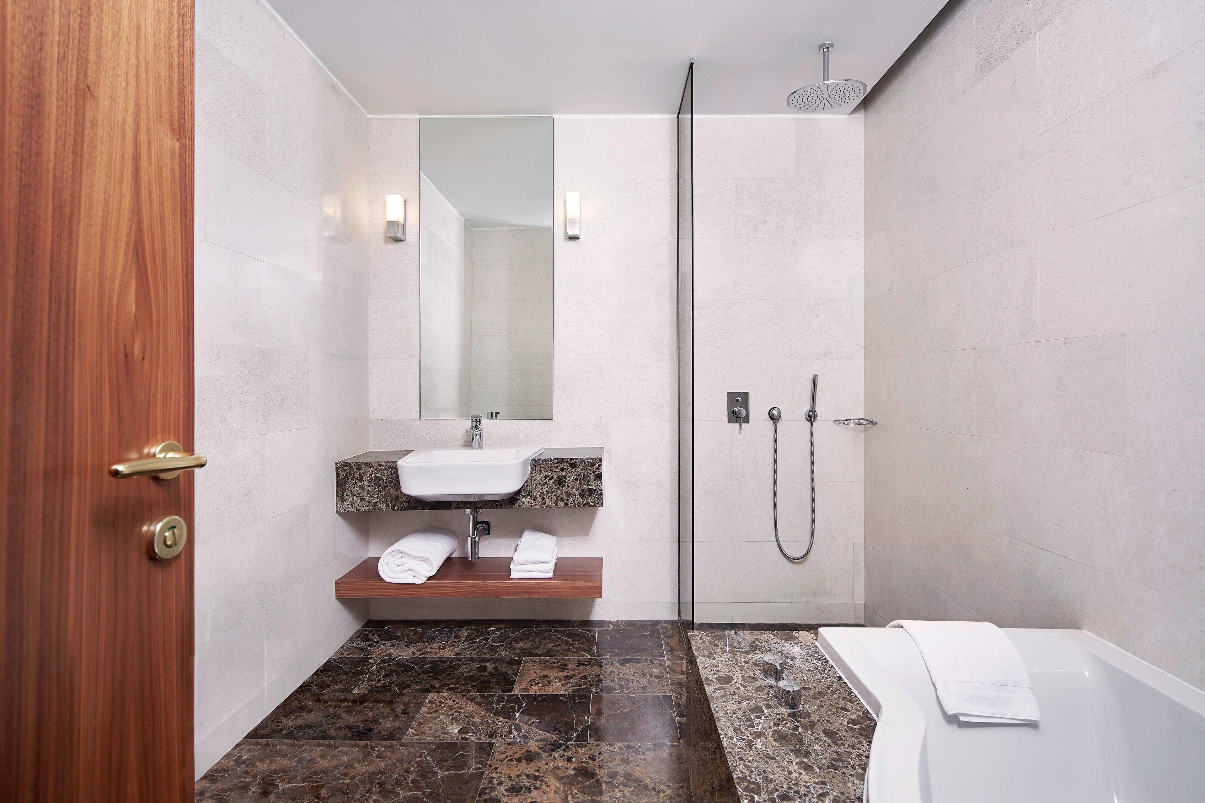 Une salle de bain moderne avec une porte en bois, un lavabo doté d’un plan de travail en marbre, un miroir, une douche avec une porte en verre et une baignoire.