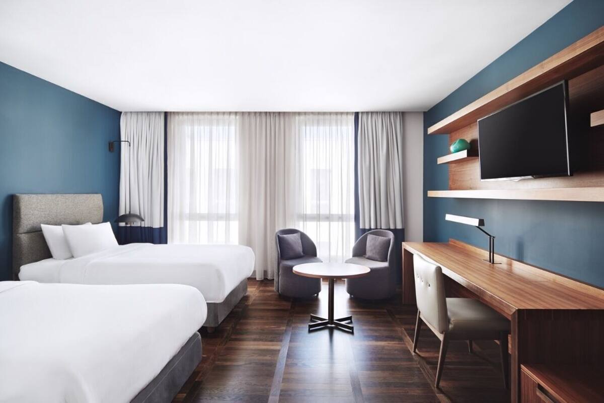 A hotel room with two beds, a desk, a chair, a TV, and a window with curtains.