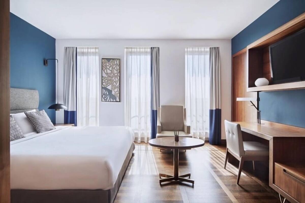 A hotel room with a bed, a round table, a chair, a desk, and a TV.