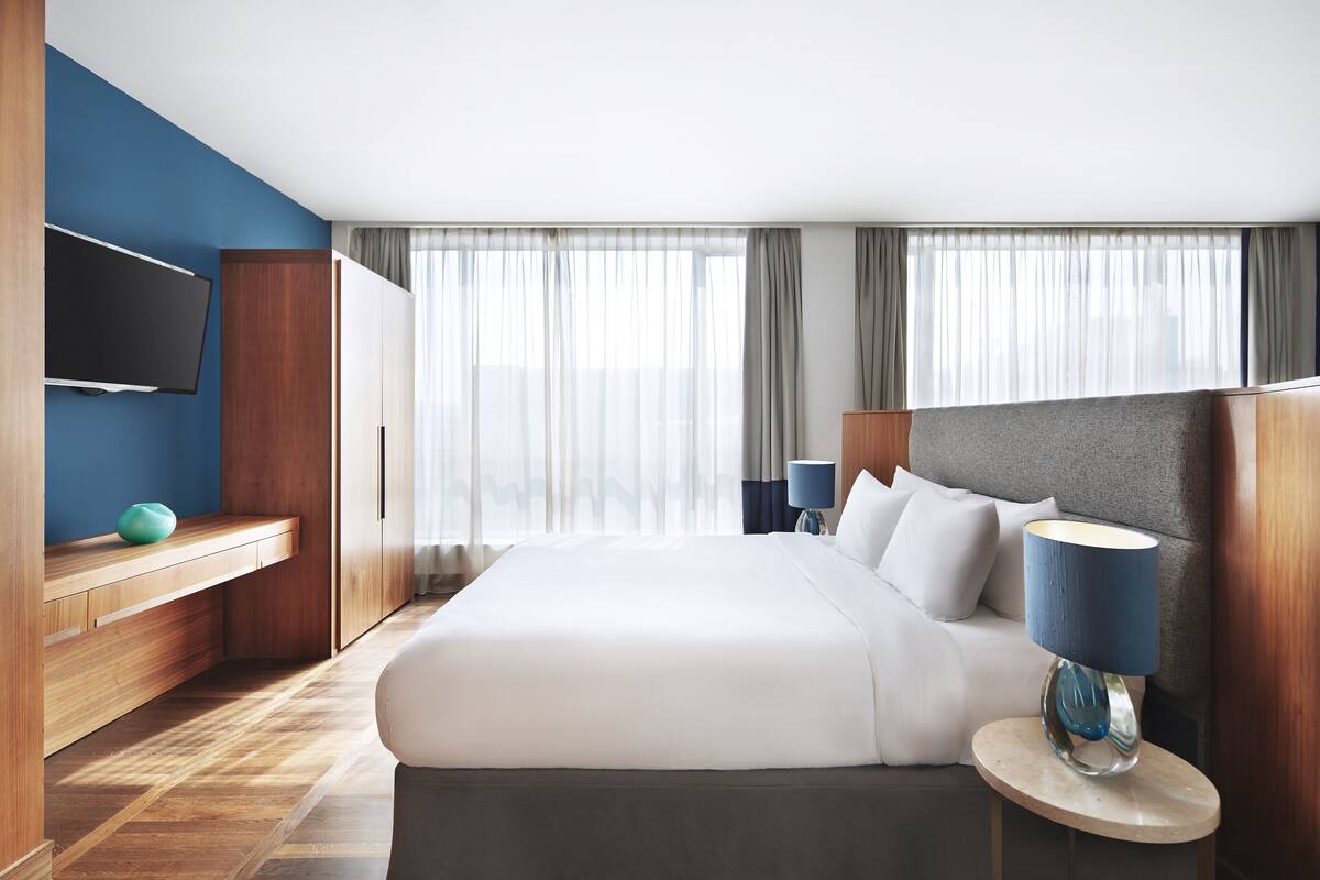 A modern hotel room with a large bed, a flat-screen TV, a wooden cabinet, and a round side table with a lamp.