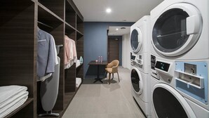 Een wasserette met twee grote wasmachines voor geladen wasgoed, een tafel met een stoel en een rek met opgevouwen kleding.