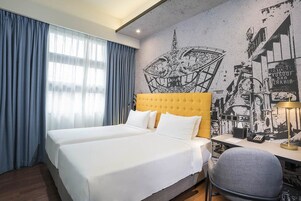 Habitación de hotel con cama doble, escritorio y silla. La habitación tiene un ventanal con cortinas, un mural en la pared y una lámpara de techo.