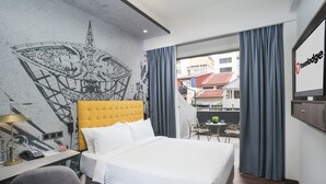Hotelkamer met een bed, een bureau met een lamp, een stoel, een televisie en een balkon met uitzicht op gebouwen.