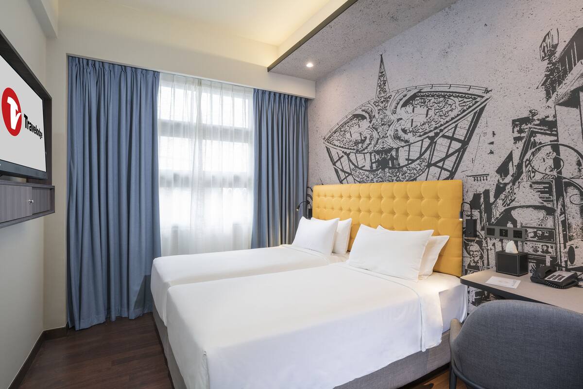Une chambre d’hôtel avec deux lits, une tête de lit jaune, un bureau avec un téléphone et une fresque murale.